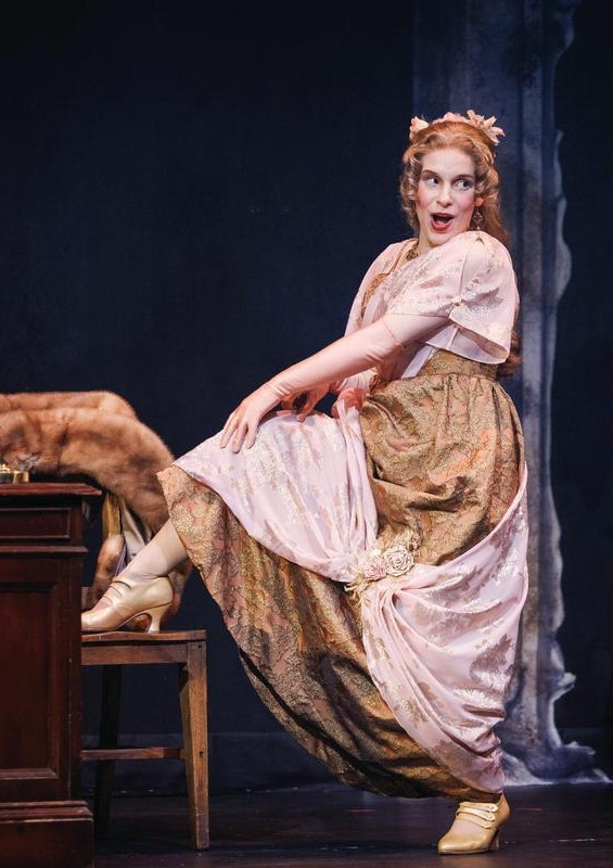 A woman in an edwardian bronze and pink gown standing with a leg on a chair