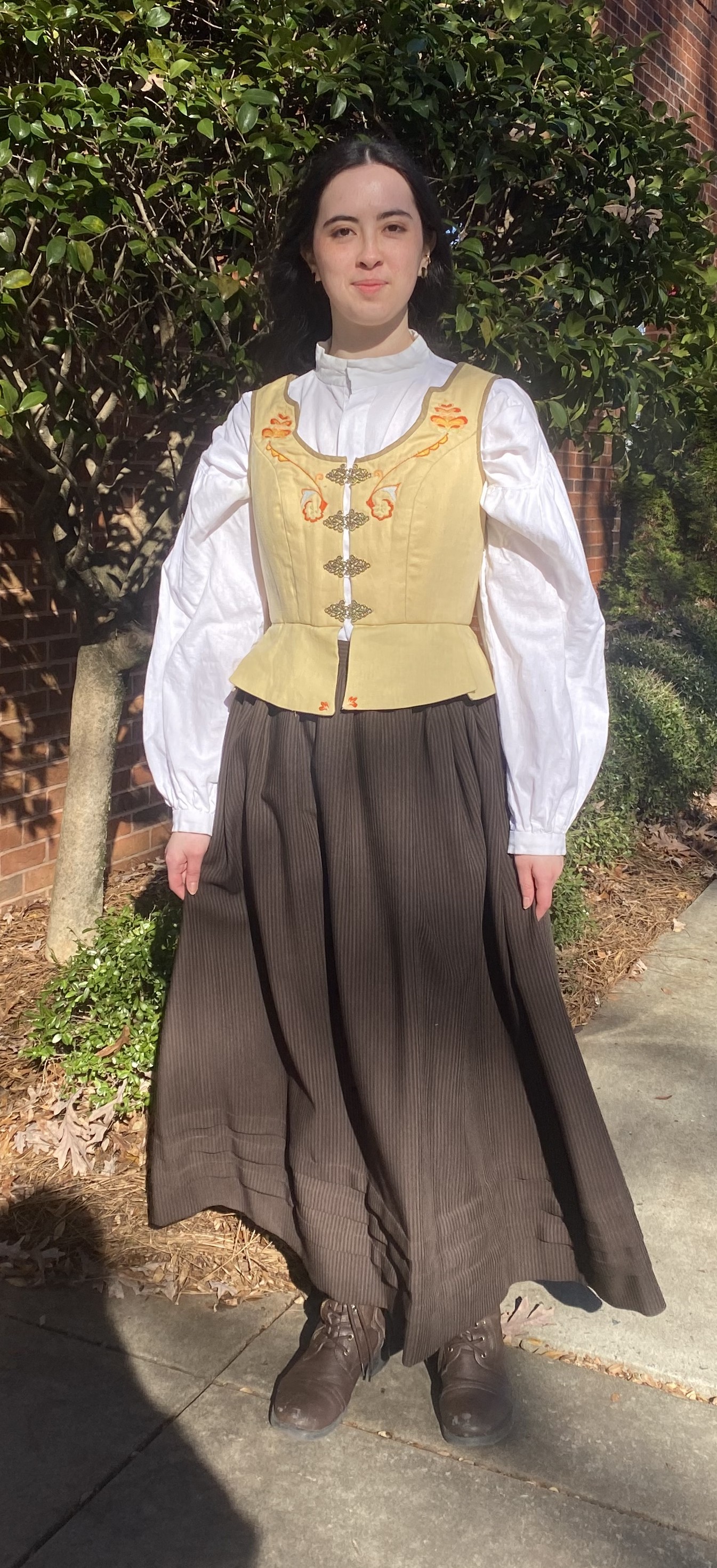 A woman in a long sleeve white shirt and long brown skirt with a yellow bodice on top, full length fron view