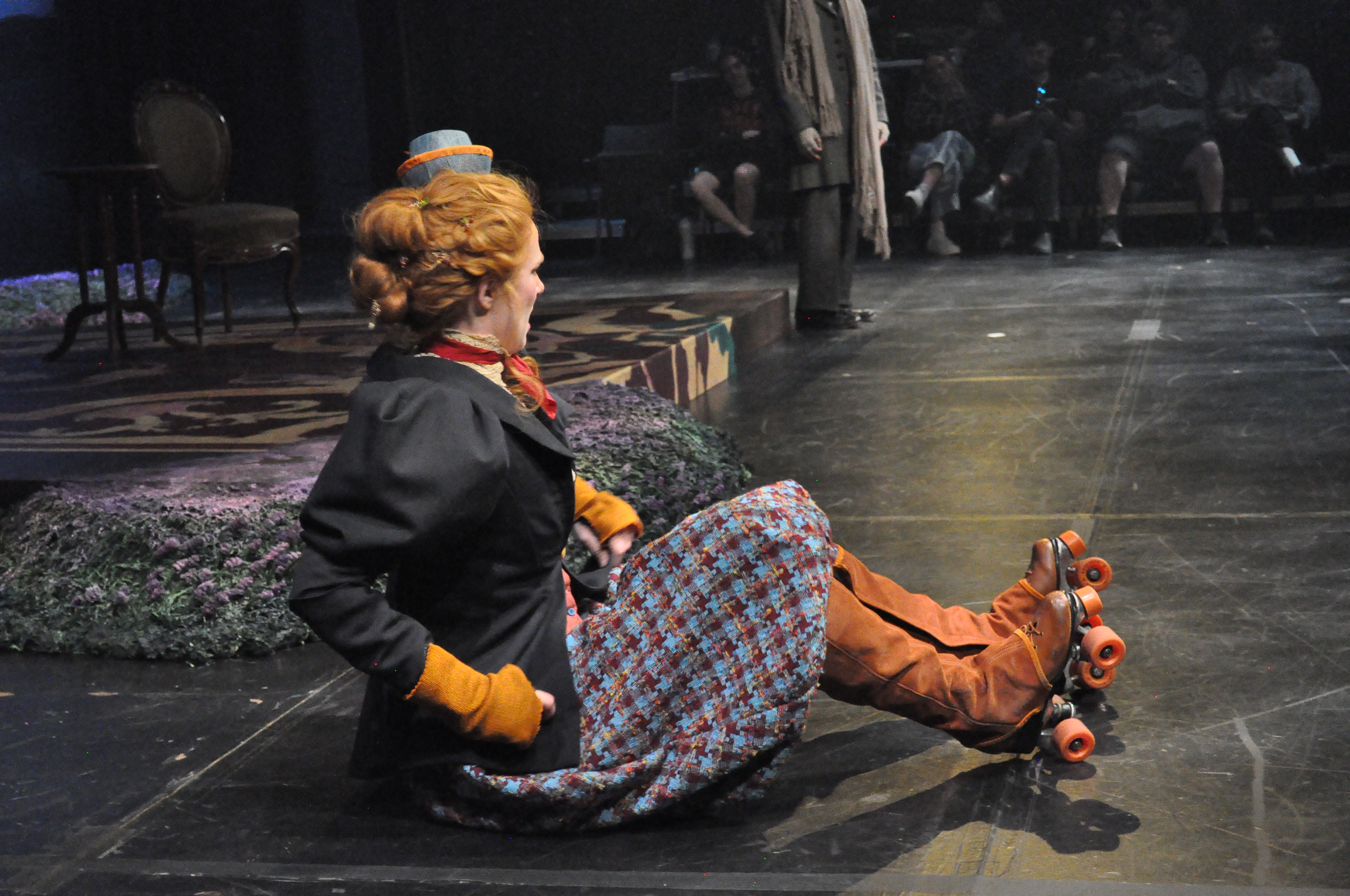 A woman on skates wearing voluminous plaid bicycle pants/breeches with a taylored vicotrian women's jacket, side view, sitting with legs out in front of her